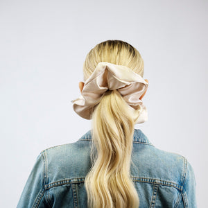 Person with long blonde hair tied in a high ponytail with a beige scrunchie, wearing a denim jacket on a white background.