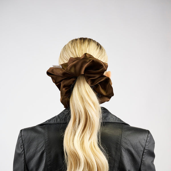 Person with long blonde hair tied in a ponytail with a brown scrunchie, wearing a black leather jacket on a white background.